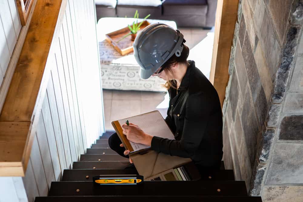 construction inspector woman sitting on the stair and taking notes using clipboard during home pre-purchase inspection,